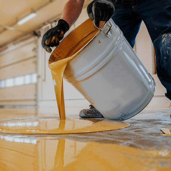 A closeup shot of a construction worker pouring out epoxy resin from a bucket onto a floor